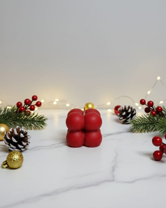 A red bubble soy wax candle with Christmas decorations like pine cones and berries on a marble surface.