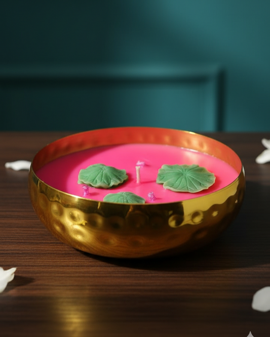 Pink candle in a gold holder with green leaves on a wooden surface against a teal wall.