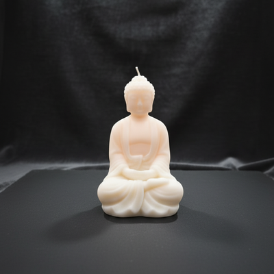 A light pink, meditating Buddha shaped soy wax candle sits on a dark surface against a black backdrop.
