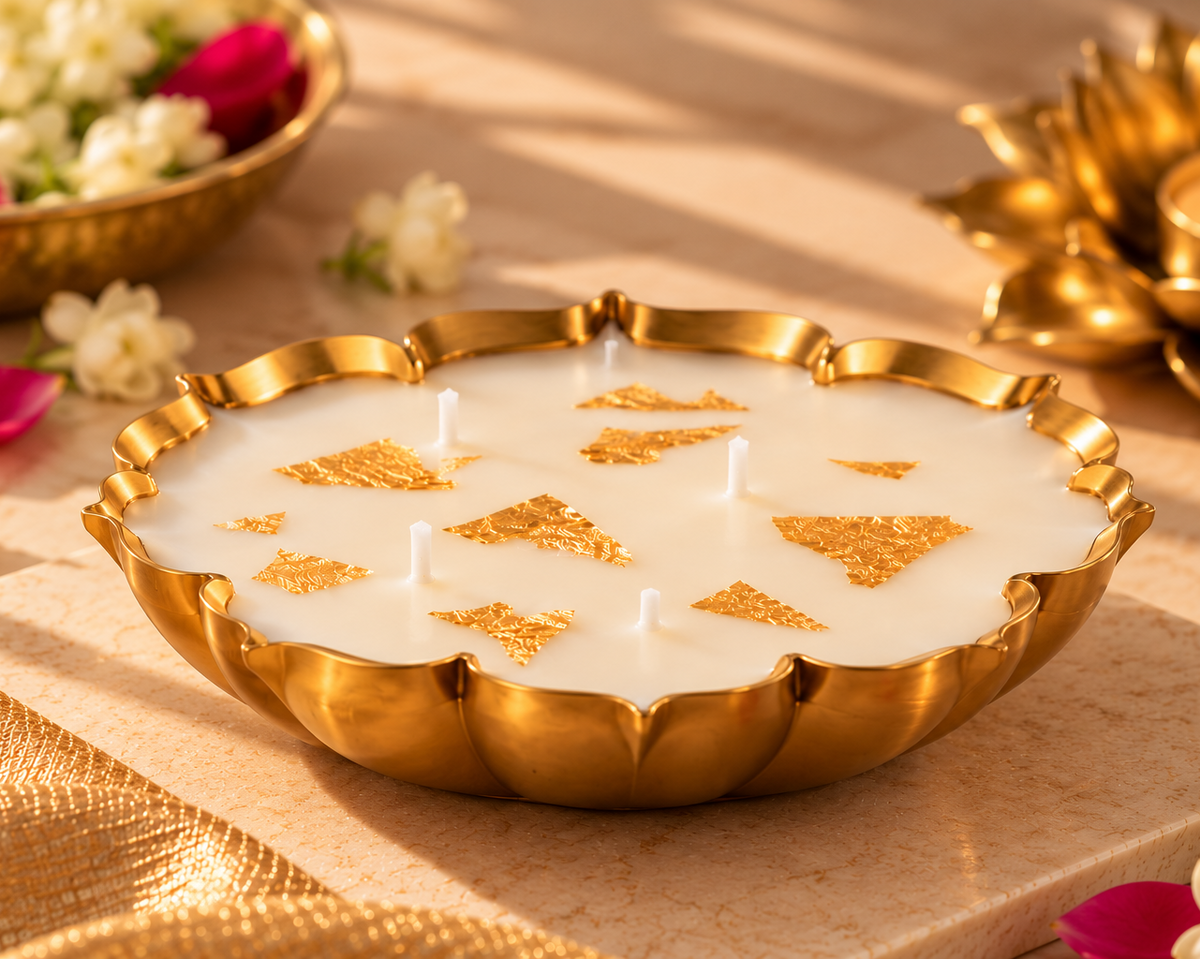 Gold scalloped urli candle with gold leaf on a marble tray beside jasmine flowers and a brass lotus diya
