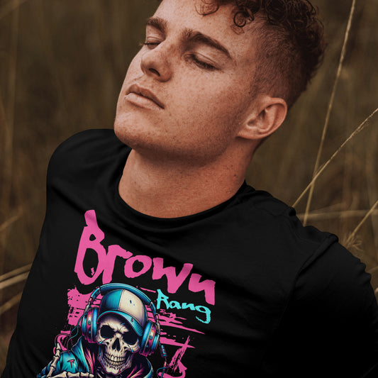 A young man with curly hair and closed eyes is wearing a black t-shirt with a vibrant graphic design. The artwork features a skeleton in a hoodie and headphones, with graffiti-style text above that reads "Brown Rang" and below "MAINE LIKHA HAI." The design has neon pink and blue accents with a street art vibe. The background shows tall grass, suggesting an outdoor setting.