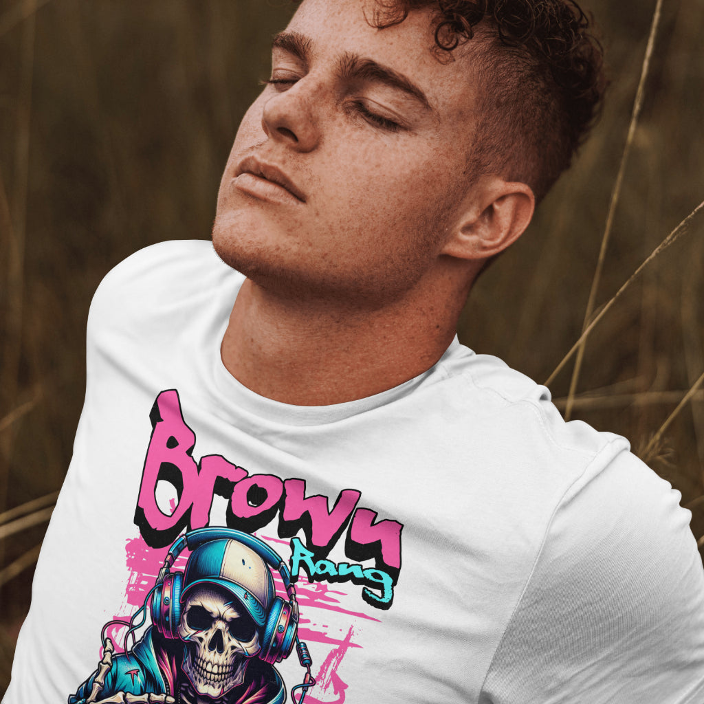 A young man with curly hair and closed eyes is wearing a white t-shirt with a vibrant graphic design. The artwork features a skeleton in a hoodie and headphones, with graffiti-style text above that reads "Browy Rang" and below "MAINE LIKHA HAI." The design has neon pink and blue accents with a street art vibe. The background shows tall grass, suggesting an outdoor setting.