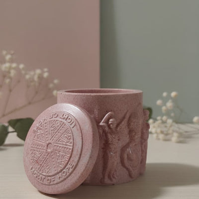 Pink Jesmonite Renaissance Relic Scrolled candle vessel with lid, featuring embossed figures and text.