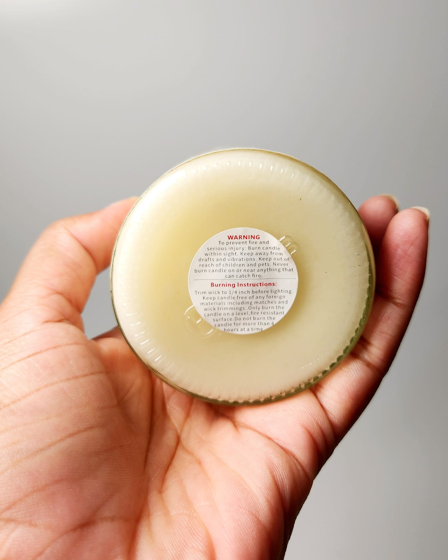 Hand holding a candle jar with a warning label against a neutral background
