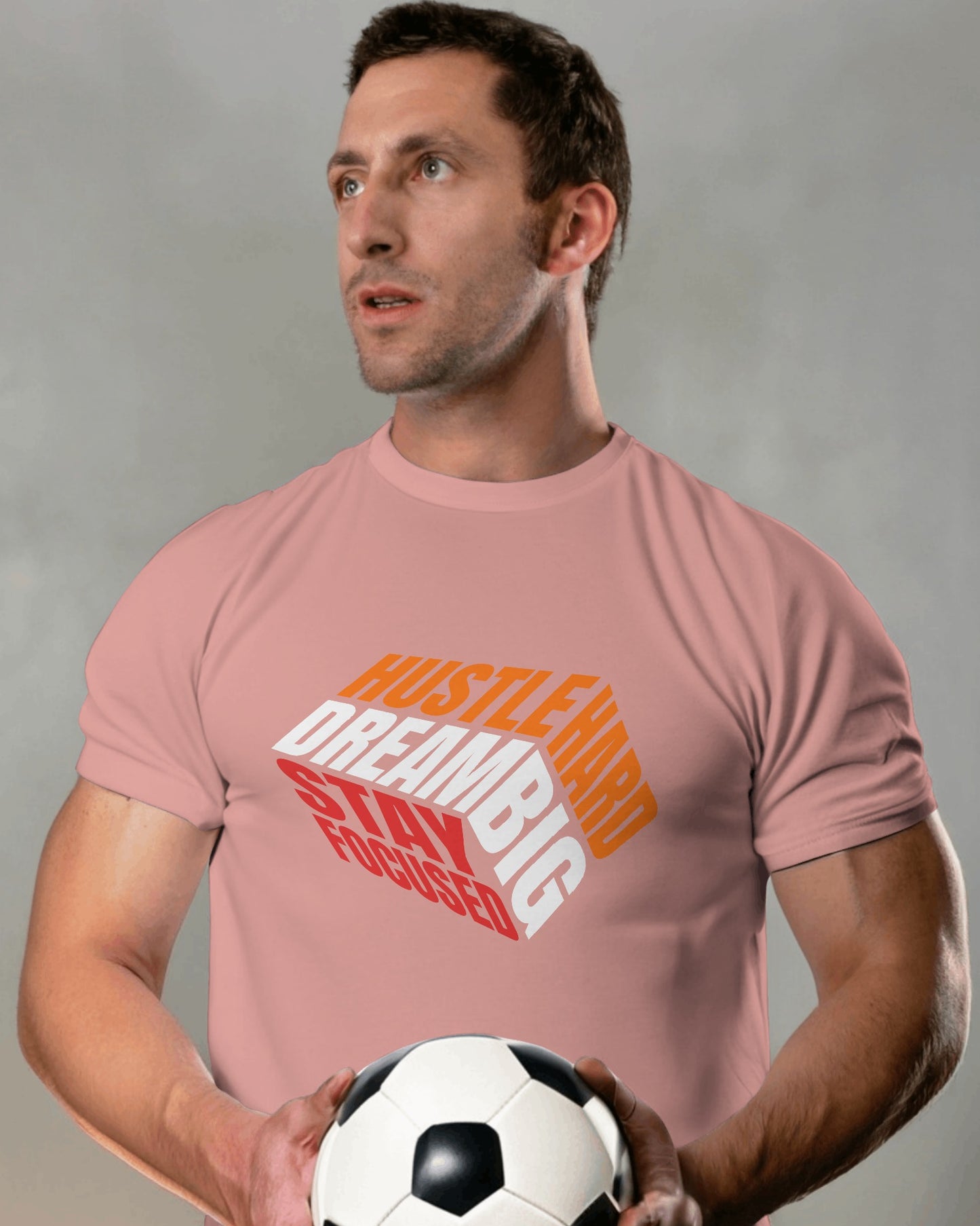 A male model standing with a football in his hands wearing 'Hustle Hard' tshirt in Flamingo color