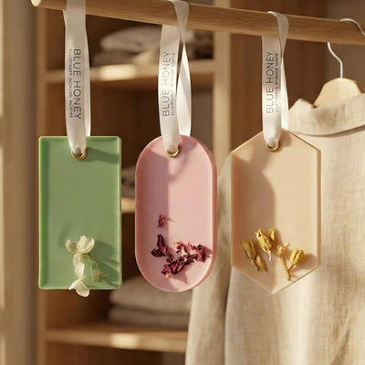 Three handcrafted natural beeswax wax sachets in green, pink, and beige with dried flowers, hanging on a wooden rod.