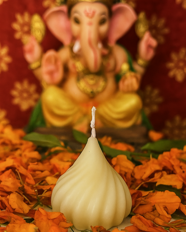 A modak-shaped ivory candle sits in front of a blurred statue of Lord Ganesha, surrounded by marigold petals.