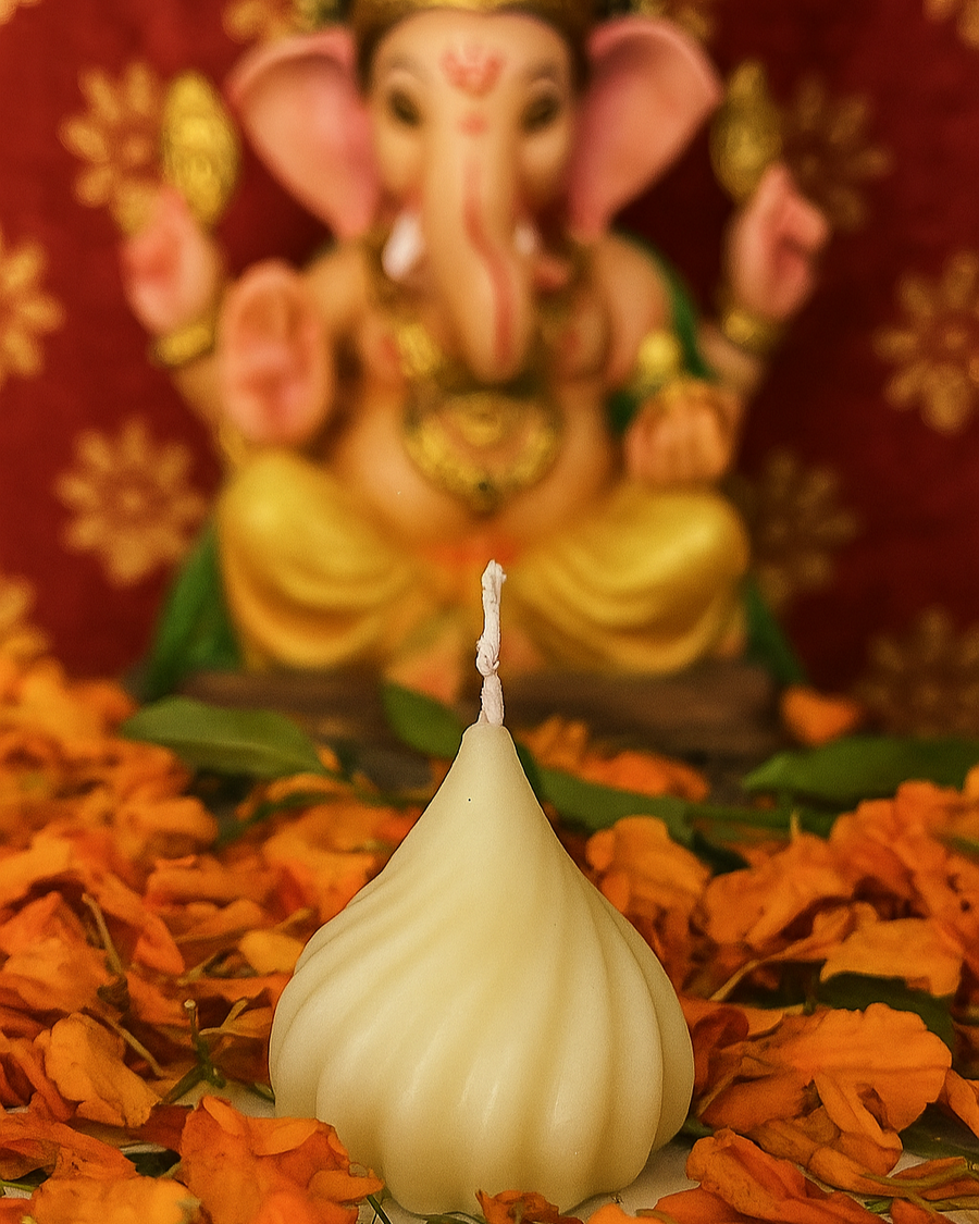 A modak-shaped ivory candle sits in front of a blurred statue of Lord Ganesha, surrounded by marigold petals.