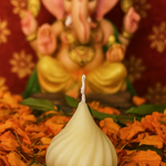 A modak-shaped ivory candle sits in front of a blurred statue of Lord Ganesha, surrounded by marigold petals.