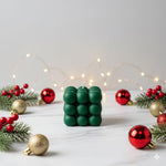 A green bubble candle sits on a marble surface surrounded by festive Christmas decorations and fairy lights.