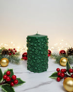 A green pillar candle with a floral vine design, decorated for Christmas with holly and baubles.