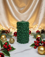 A green pillar candle with a floral vine design, decorated for Christmas with ornaments and holly.