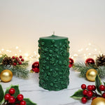A green pillar candle with a floral vine design, decorated for Christmas with holly and baubles.