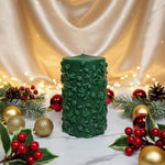 A green pillar candle with a floral vine design, decorated for Christmas with ornaments and holly.
