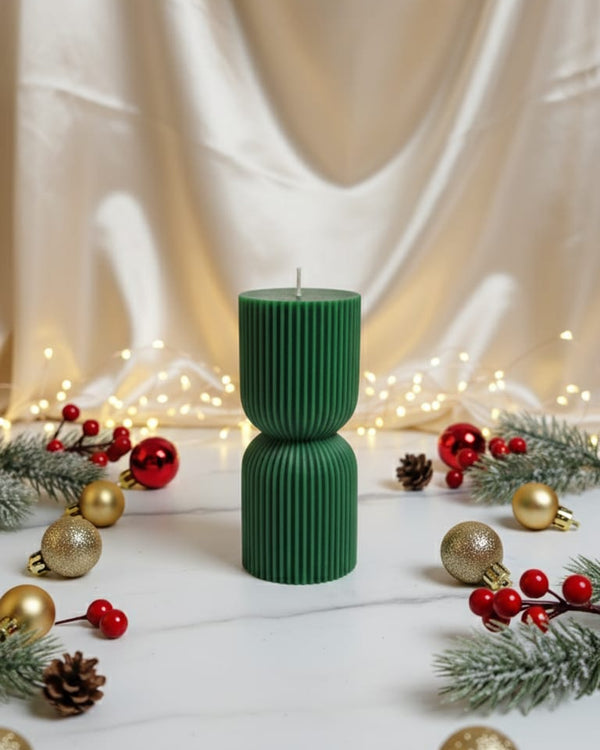 A festive green hourglass pillar candle surrounded by red berries, gold ornaments, and pine sprigs on a marble surface.