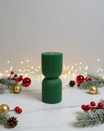 A green hourglass-shaped soy wax candle with ribbed texture, decorated for Christmas with pine sprigs and ornaments.