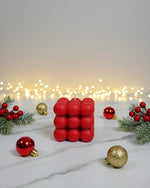 A red bubble candle sits on a marble surface surrounded by festive Christmas decorations and fairy lights.