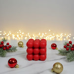A red bubble candle sits on a marble surface surrounded by festive Christmas decorations and fairy lights.