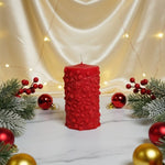 A red pillar soy wax candle with a floral pattern, decorated for Christmas with ornaments and pine branches.