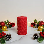 A red Christmas pillar candle with floral embossing, surrounded by festive decorations like holly and baubles.
