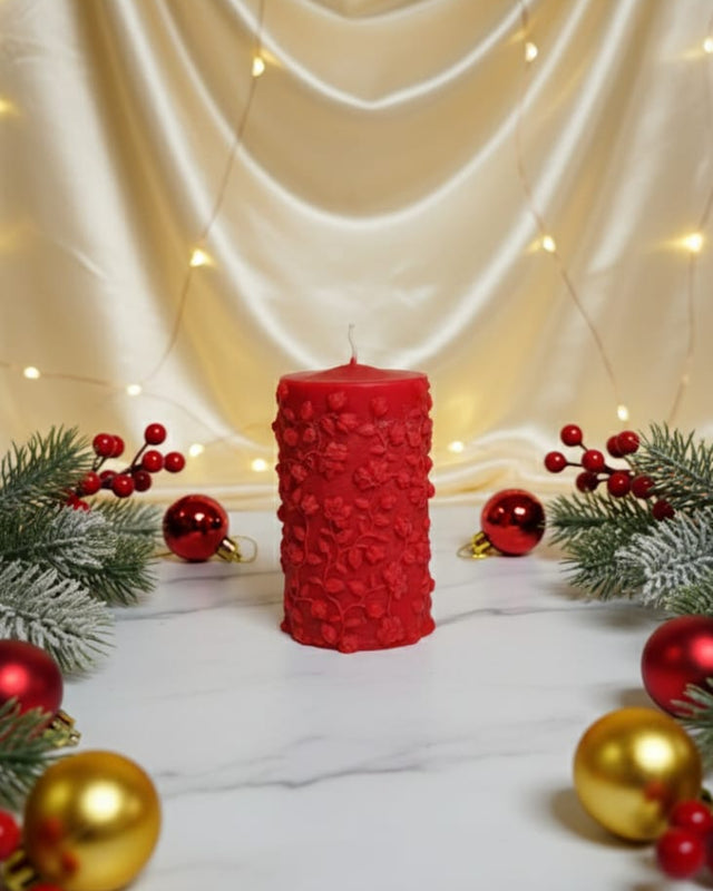 A red pillar soy wax candle with a floral pattern, decorated for Christmas with ornaments and pine branches.