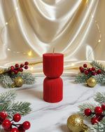 A red hourglass-shaped pillar candle with ribbed texture, decorated for Christmas with holly and baubles.