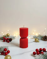 A red hourglass-shaped pillar soy wax candle with a ribbed texture, set against a festive Christmas background.