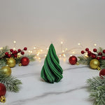 A green, twisted Christmas tree-shaped soy wax candle sits on a marble surface, surrounded by festive decorations.
