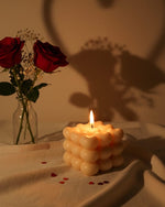 A lit sculpted bubble candle with a heart-shaped shadow, next to red roses in a vase.