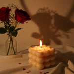 A lit sculpted bubble candle with a heart-shaped shadow, next to red roses in a vase.