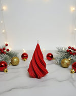 A red, twisted Christmas tree-shaped soy wax candle sits on a marble surface surrounded by festive ornaments and greenery.