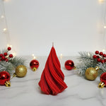A red, twisted Christmas tree-shaped soy wax candle sits on a marble surface surrounded by festive ornaments and greenery.