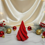 A red, twisted Christmas tree-shaped soy wax candle sits on a marble surface with festive decorations.