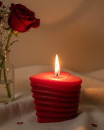 A red heart-shaped sculpted candle with a lit wick, next to a red rose in a vase.