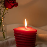 A red heart-shaped sculpted candle with a lit wick, next to a red rose in a vase.