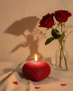 A red heart-shaped sculpted candle with a lit flame, beside two red roses in a vase.