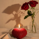 A red heart-shaped sculpted candle with a lit flame, beside two red roses in a vase.