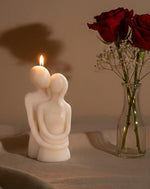 A lit sculpted candle of a couple embracing, with red roses in a vase in the background.