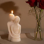 A lit sculpted candle of a couple embracing, with red roses in a vase in the background.