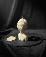 Ivory blindfolded oracle bust candle with floral accents, displayed on a dark slate with a leaf and stone.