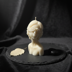 Ivory blindfolded oracle bust candle with floral accents, displayed on a dark slate with a leaf and stone.