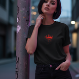 A woman wearing a black 'Wear the Crown. Own the Hype' t-shirt with a red crown graphic, leaning against a pole.