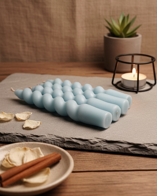 Blue candle with a textured design on a wooden surface with a plant and candle holder in the background.
