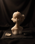 White scented candle in the shape of a maiden's bust with floral accents, presented on a dark slate.