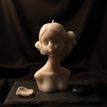 White scented candle in the shape of a maiden's bust with floral accents, presented on a dark slate.