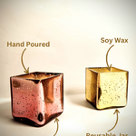 Two scented cuboid-shaped glass jar candles, one gold and one rose gold, with labels indicating hand-poured, soy wax, and reusable jar.