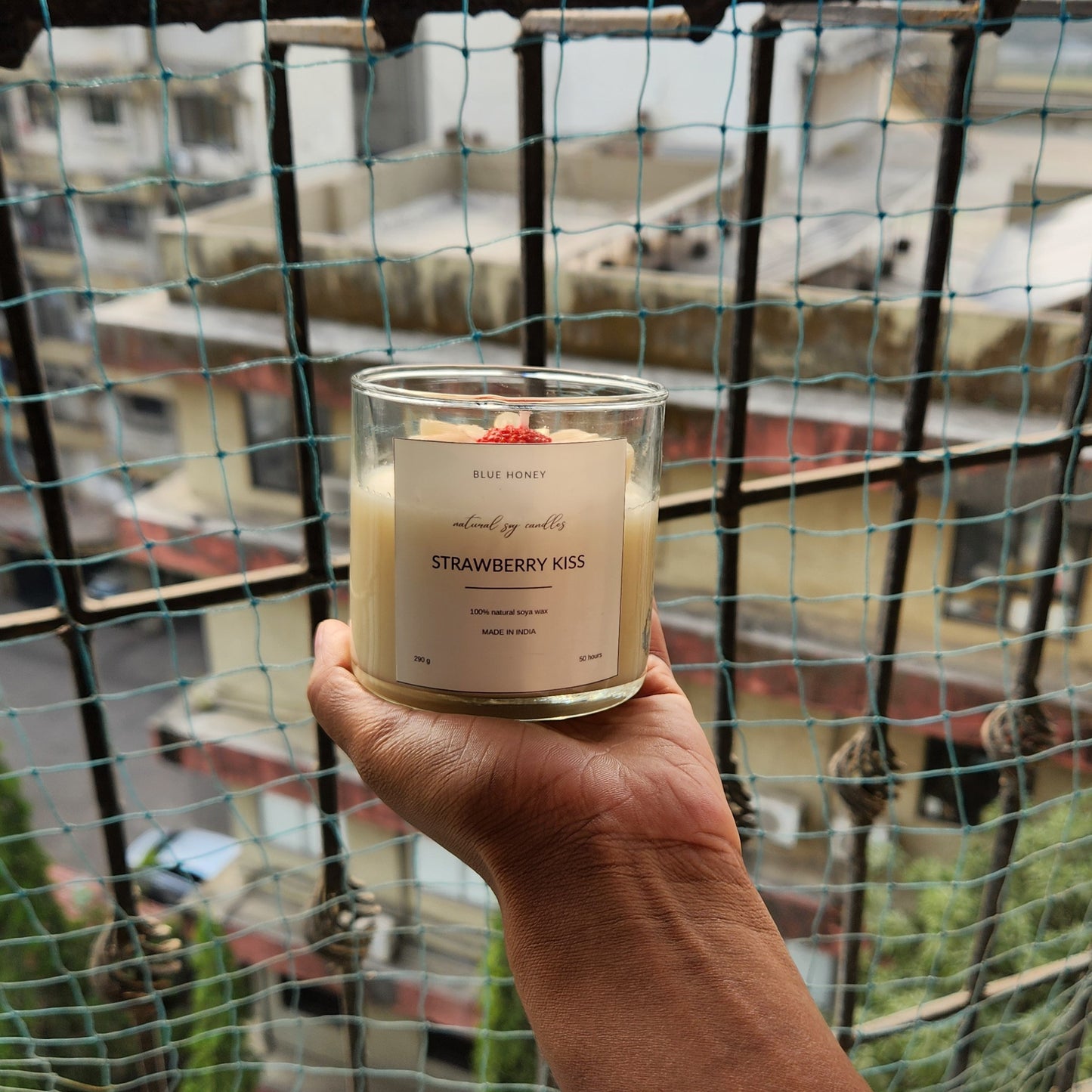 Red bud daisy flower candle inside a clear jar in palm of a female, outside the window. label reads Strawberry Kiss.