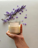 A hand holds a clear glass soy wax candle labeled 'Lavender Bliss' with lavender sprigs and scattered petals.