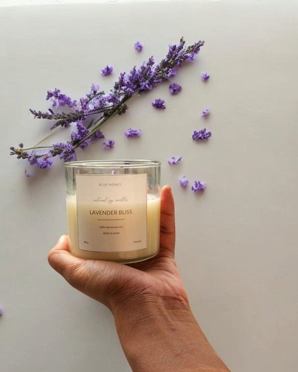 A hand holds a clear glass soy wax candle labeled 'Lavender Bliss' with lavender sprigs and scattered petals.