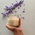 A hand holds a clear glass soy wax candle labeled 'Lavender Bliss' with lavender sprigs and scattered petals.